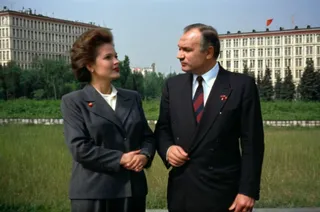 Two Soviet politicians from the 1980s, a woman and a man, dressed in formal suits, standing outdoors with socialist Soviet housing buildings and greenery in the background.