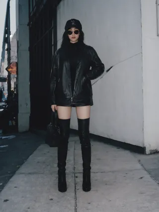 Full body portrait of a woman modeling a black leather outfit with over-the-knee boots, black sunglasses, and a cap on the streets of New York.