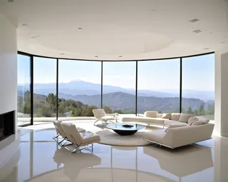 Bright modern living room with white furniture, floor-to-ceiling windows showcasing a panoramic mountain view, glossy white flooring, and an elegant circular fireplace.