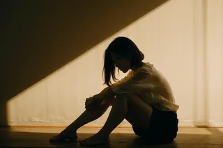 Silhouette of a mature woman sitting on the floor with knees bent, leaning forward, illuminated by evocative shadows and nuanced chiaroscuro lighting.