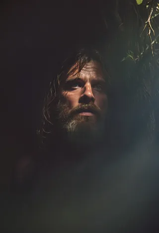 Close-up portrait of a bearded man with wet hair and intense eyes, illuminated by dramatic glimmering light with dark shadows surrounding him