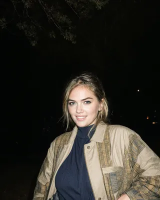 Portrait of a smiling woman with blonde hair wearing a plaid coat and navy turtleneck, set against a dark nighttime background with soft lighting.