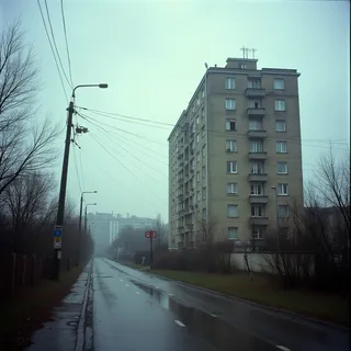1990s Soviet Russia street scene with a tall apartment building on the right, overcast sky, wet empty road, and leafless trees creating a gloomy atmosphere