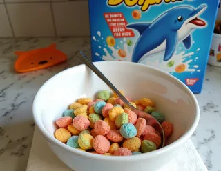 Biała ceramiczna miska wypełniona kolorowymi, okrągłymi płatkami z łyżką, umieszczona na serwetce na kwarcowym blacie, z kolorowym pudełkiem Dolphin Cereal przedstawiającym szczęśliwego delfina wyskakującego z mleka w tle.