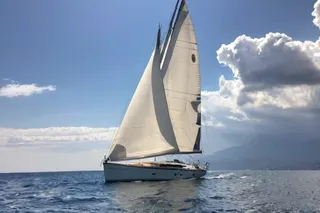 A white sail boat with sails fully raised sailing on calm blue water near the Italian coastline under a bright blue sky with fluffy clouds.