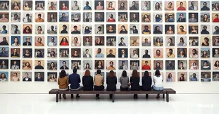 Group of people seated on a bench facing a large white wall covered with diverse portrait photographs arranged in a mosaic pattern, viewed from behind under natural light.