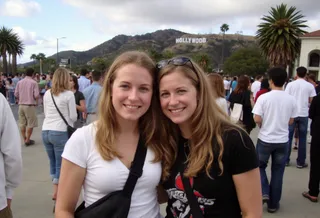 Two blonde sisters in casual clothing smiling and posing on a crowded street with the Hollywood sign visible on the hillside behind them in Los Angeles.
