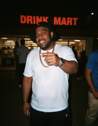 Smiling man in white t-shirt and black cap with gold chain standing outside Drink Mart convenience store at night