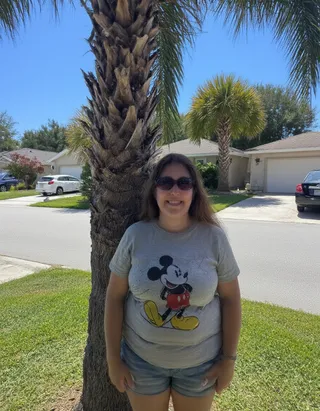 Female tourist wearing a grey Mickey Mouse t-shirt and denim shorts, standing and smiling in front of a palm tree in a sunny suburban Florida neighborhood.
