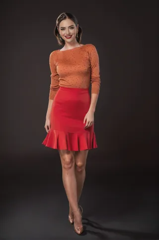 A realistic full-body image of a smiling woman with brown hair wearing a red dress, brown top, earrings, makeup, and pantyhose standing against a dark background.