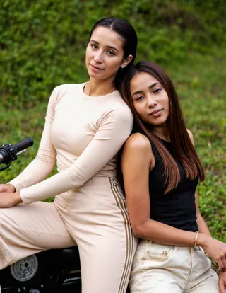 Two women sitting together on a motorcycle in an outdoor setting, wearing neutral-toned casual outfits with a green natural background.