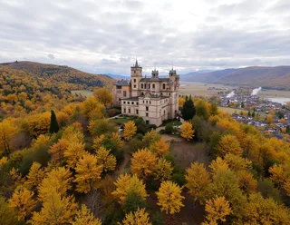 Aerial view of a European countryside castle surrounded by vibrant autumn trees with golden-yellow leaves, rolling hills, and a distant town under a cloudy sky.