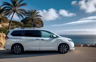 White Nissan Quest minivan parked on a coastal road with California cliffs, palm trees, an ocean beach, and a partly cloudy sky at sunset.
