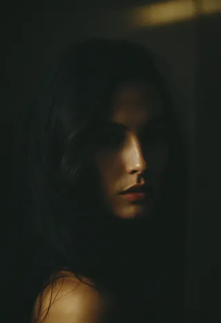 Low key sensual portrait of a mysterious woman with soft lighting highlighting her face and shoulder against a dark background.