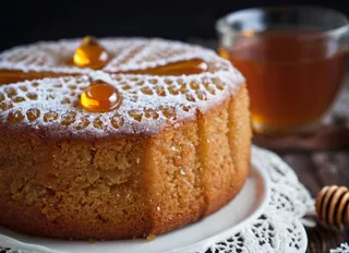 Close-up of a Russian Honey cake decorated with powdered sugar and honey drops on top, served on a white plate with lace doily, with a glass of tea in the background.