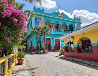 A vibrant Caribbean city street shows a turquoise government building with revolutionary murals, surrounded by palm trees and colorful tropical flowers under a bright blue sky.