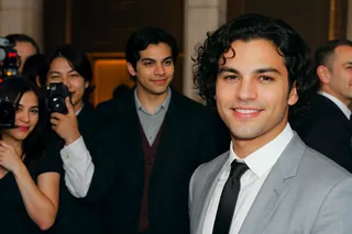 A smiling man with curly black hair wearing a light gray suit and black tie at a red carpet event, with photographers and a sponsorship poster visible in the background.