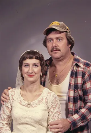 Vintage 1979 bridal portrait showing a bride in lace wedding dress with heavy makeup and a groom wearing a trucker cap, flannel shirt, and white tank top.
