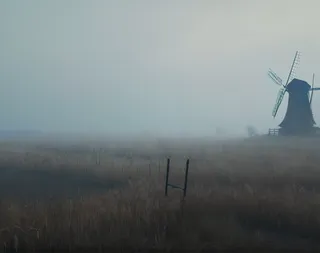 A foggy early winter landscape in Eastern Europe featuring a dying grass field, a small village on the left, and an old windmill on the right in cold, windy weather.