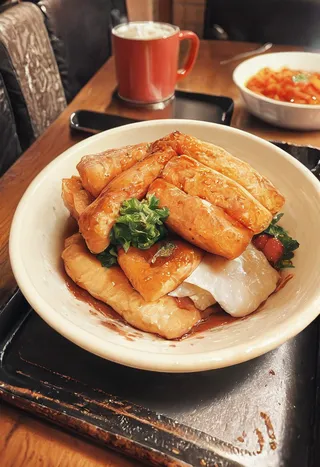 Close-up of fried tofu served in a white bowl with greens, placed on a rustic tray in a cozy restaurant setting with warm lighting and a red mug in the background.