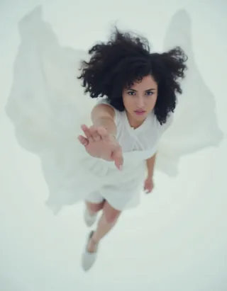 Top view of a woman with black curly hair falling, wearing a flowing white dress, reaching her hand toward the viewer with a blurred, cinematic effect.