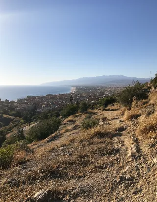 A scenic view showing a roman city nestled in a hilly Italian landscape with ocean on the left and mountains in the background, captured in an iphone photo style.
