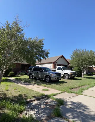 Daytime photo of a Texan suburban neighborhood featuring single family homes, neatly maintained lawns, a parked SUV, and a white pickup truck under a clear blue sky.