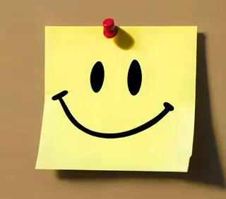 Yellow sticky note pinned with a red pin featuring a black smiley face drawn on it.