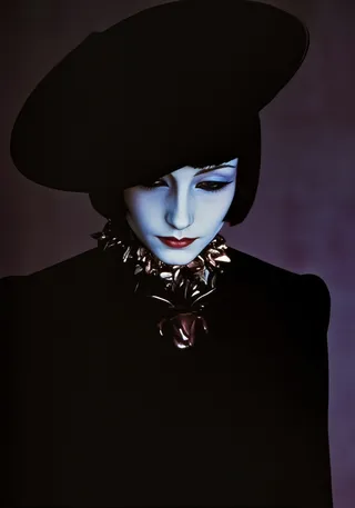 Portrait of a woman with pale skin wearing a large black hat, dramatic dark clothing, and an ornate antique necklace, looking downward with red lips and dark eye makeup.