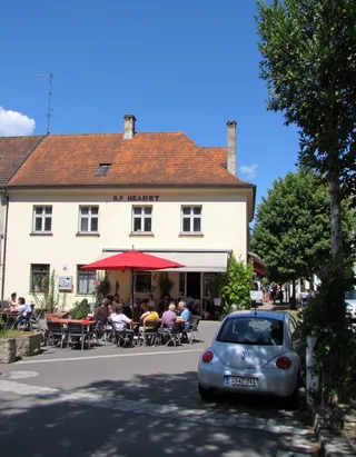 Sunny summer day at a cafe in a small German town featuring outdoor seating with people, a parked silver VW car with a European license plate, and a clear blue sky.