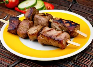 Grilled shish kebab skewers with juicy pieces of meat on a yellow and white plate accompanied by sliced cucumber and tomato in the background