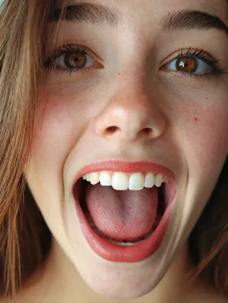 Close-up of a young woman with brown eyes and long hair, mouth wide open displaying perfect and symmetric teeth, detailed skin texture, and visible tongue.