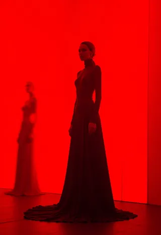 High fashion photograph of two women standing in front of a bright red wall with motion blur effect and non-centered composition, showcasing elegant long dresses.