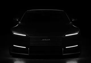 Front view of a sleek black Jaguar Type car in a dark studio with LED headlights and illuminated horizontal grille lines, highlighting its glossy paint and chrome accents.