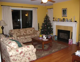 Living room interior from the 2000s featuring floral patterned sofas, a decorated Christmas tree in the corner, yellow wallpaper, a fireplace with mantle decor, a wooden coffee table, and clutter.