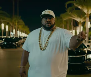 Obese black man wearing a white t-shirt and white cap, adorned with large gold chains, pointing while standing on a night-time street lined with palm trees and street lamps, with a green Chrysler 300 low rider car in the background.