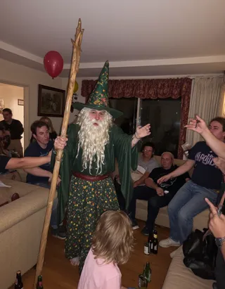 A person dressed as a wizard with a long white beard, floral pointy hat and robe, holding a wooden staff, in the middle of a casual party in a living room with guests seated on sofas and balloons in the background.