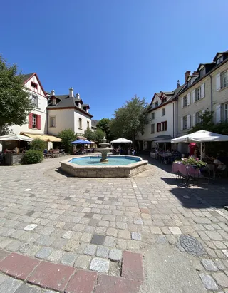 Sunny French village square with an octagonal stone fountain in the center, surrounded by cobblestone roads, white and beige houses, market stalls with umbrellas, and villagers enjoying a summer day under clear blue sky.