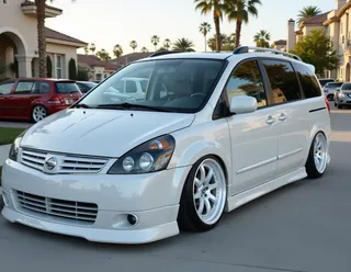 White Nissan Quest minivan with tinted windows, low suspension, enlarged grille, white wheels, and chrome details parked on a suburban street.