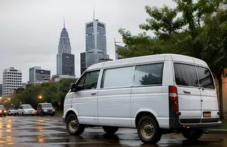 Old white van parked on a wet city street with reflections and an urban skyline featuring tall skyscrapers under a cloudy sky.