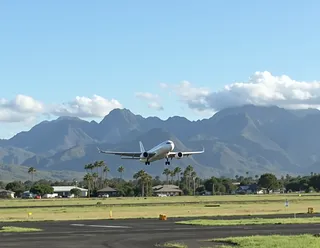 Airplane landing on an airport runway with lush palm trees and majestic Hawaiian mountains in the background under a clear blue sky.