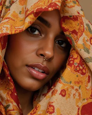 Close-up portrait of a woman with warm brown skin partially covered by a brightly colored scarf featuring intricate red, orange, and yellow floral and paisley patterns, highlighting her expressive eyes and nose ring under soft warm lighting.