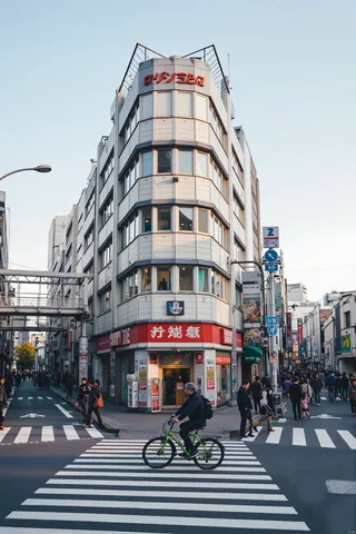 A man on a green bicycle crossing a city street crosswalk with a white multi-story building with red signage in the background and people walking nearby under a clear blue sky.