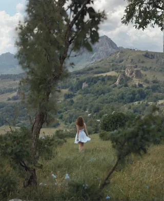 A woman in a white dress walking away through a green meadow surrounded by trees toward mountainous terrain under a cloudy sky.