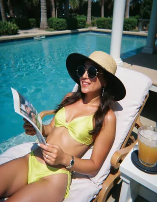 A woman in a bright yellow bikini reclines on a white lounger beside a clear blue pool. She wears a wide-brimmed straw hat and round sunglasses while reading a magazine on a sunny summer day.