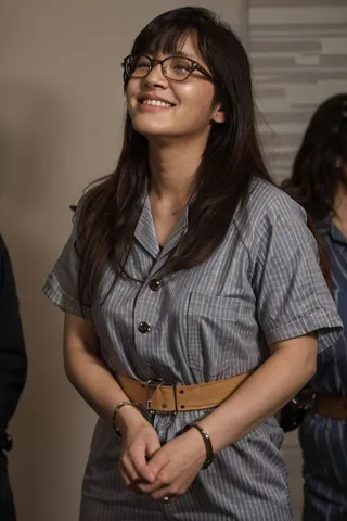 Smiling Asian teen girl wearing rectangle glasses and a striped prison jumpsuit with handcuffs and a belt, in a courtroom setting.