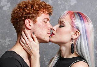 Close-up of a goth girl with pastel pink, lavender, and gray hair kissing a freckled young man with curly red hair, both expressing tenderness and affection.