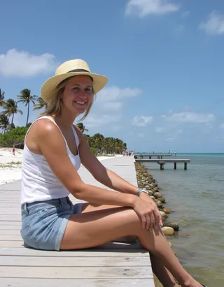 A young woman sitting on a wooden pier at a Florida beach, wearing a white tank top, jean shorts, and a sun hat, smiling under a sunny sky with palm trees in the background.