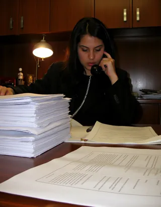 A stressed lawyer with black hair in a black suit sits behind a desk cluttered with large stacks of documents, speaking on a corded phone in a dimly lit office.