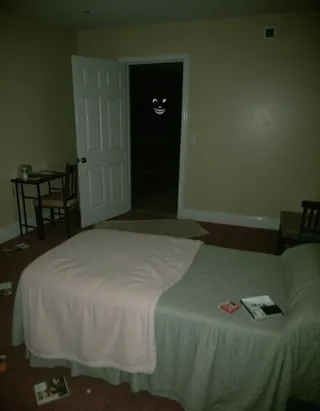 Low quality grainy photo of a creepy abandoned bedroom with a messy bed and an open door revealing a dark space with a slight visible smiling face.
